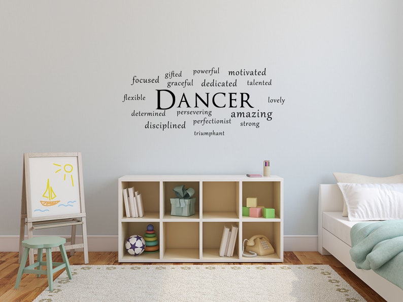 May include: A white wall with a black vinyl decal that reads "DANCER" surrounded by positive words describing a dancer. The decal is in a child's bedroom with a white bookshelf, a white easel with a drawing of a boat and sun, a green stool, and a white bed with a blue blanket.