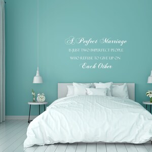 May include: A white wall with a teal blue paint color and a white vinyl wall decal with the text "A Perfect Marriage is just two imperfect people who refuse to give up on Each Other". The wall is behind a white bed with white bedding and a white nightstand with a clock and flowers.
