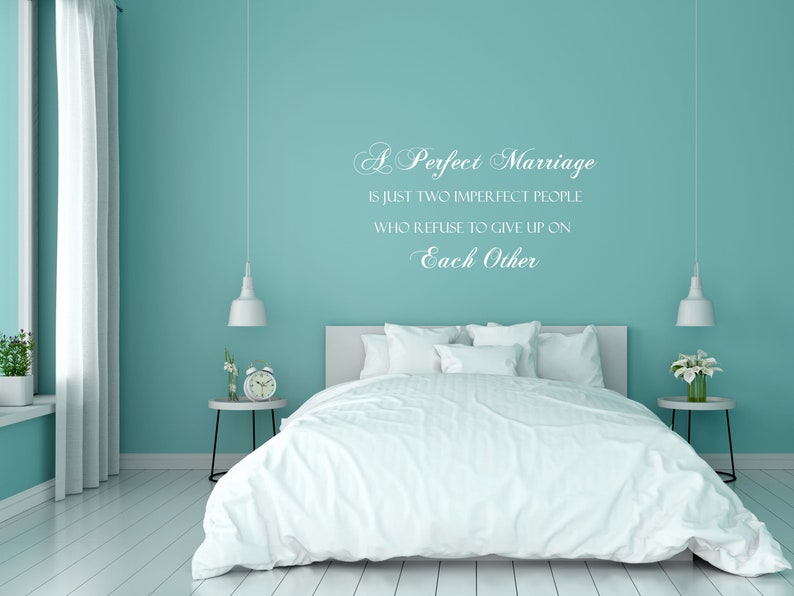 May include: A white wall with a teal blue paint color and a white vinyl wall decal with the text "A Perfect Marriage is just two imperfect people who refuse to give up on Each Other". The wall is behind a white bed with white bedding and a white nightstand with a clock and flowers.