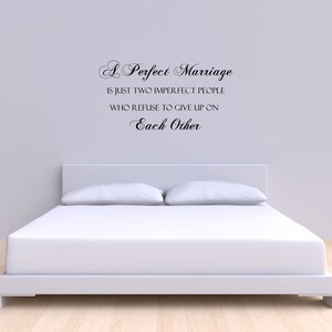 May include: A white bed with white bedding and two pillows. The wall behind the bed has a black vinyl wall decal with the text "A Perfect Marriage is just two imperfect people who refuse to give up on Each Other."