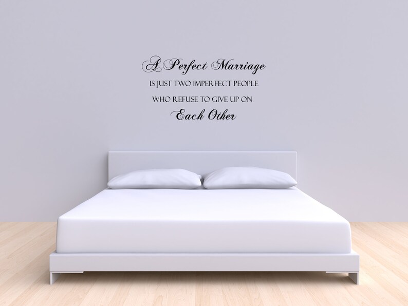 May include: A white bed with white bedding and two pillows. The wall behind the bed has a black vinyl wall decal with the text "A Perfect Marriage is just two imperfect people who refuse to give up on Each Other."