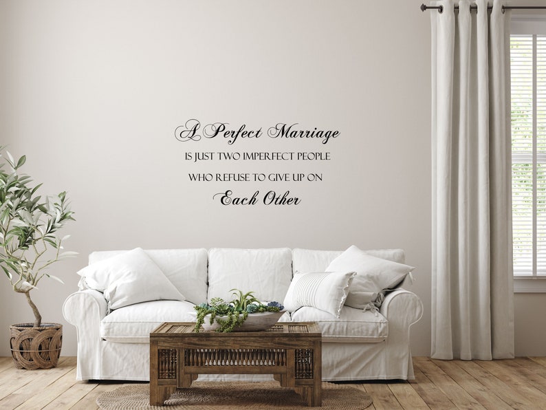 May include: A white couch with pillows in a living room setting. The wall behind the couch has a black vinyl decal with the text "A Perfect Marriage is just two imperfect people who refuse to give up on Each Other."