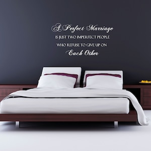 May include: A white wall with a quote about marriage written in cursive script. The quote reads "A Perfect Marriage is just two imperfect people who refuse to give up on Each Other". A bed with white linens and a wooden headboard is in the foreground.