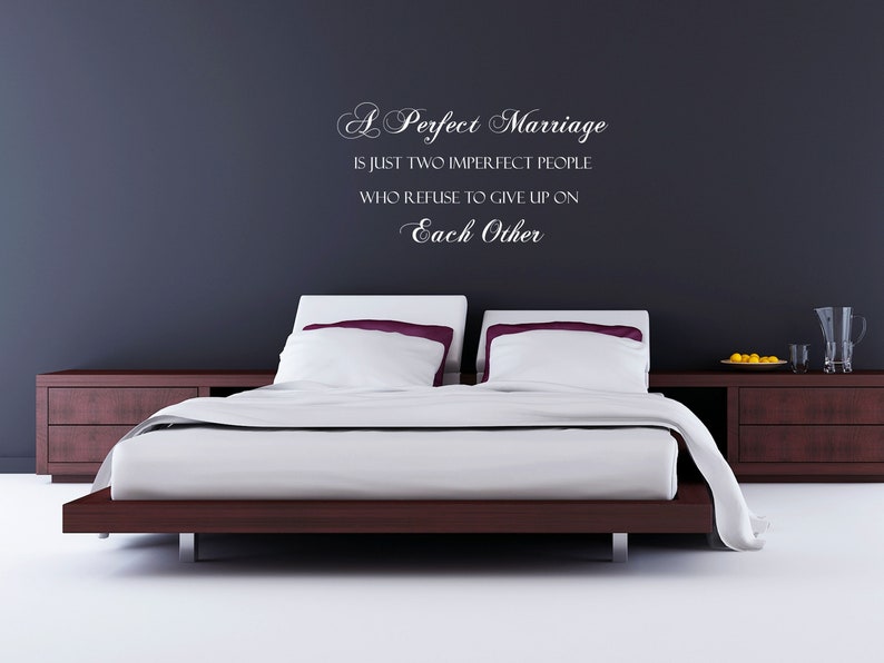 May include: A white wall with a quote about marriage written in cursive script. The quote reads "A Perfect Marriage is just two imperfect people who refuse to give up on Each Other". A bed with white linens and a wooden headboard is in the foreground.