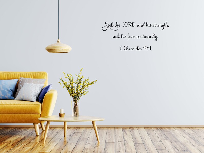 May include: A yellow couch with pillows in a living room setting with a wooden coffee table and a vase of yellow flowers. The wall has a black and white wall decal with the text "Seek the LORD and his strength, seek his face continually. I Chronicles 16-11".