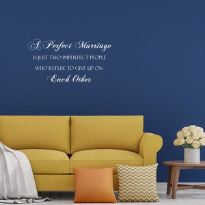 May include: A yellow couch with white and orange throw pillows in front of a blue wall with a white vinyl quote that reads "A Perfect Marriage is just two imperfect people who refuse to give up on Each Other."