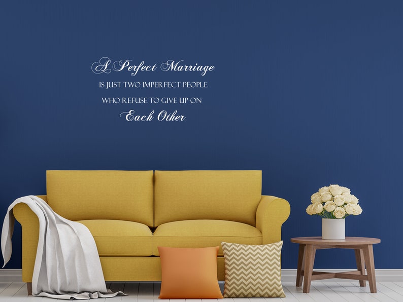 May include: A yellow couch with white and orange throw pillows in front of a blue wall with a white vinyl quote that reads "A Perfect Marriage is just two imperfect people who refuse to give up on Each Other."