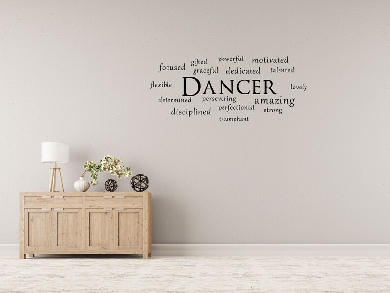 May include: A white wall with a black word cloud that reads "gifted powerful motivated focused graceful dedicated talented flexible DANCER lovely determined persevering amazing disciplined perfectionist strong triumphant". A wooden dresser with three drawers and four doors is in the foreground.