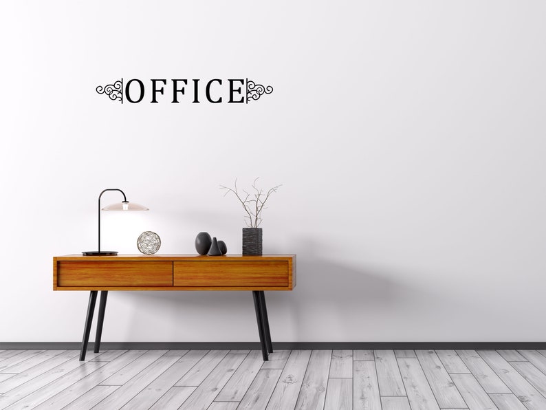 Puede incluir: Una consola de madera con dos cajones y patas de metal negro. La mesa est&aacute; decorada con una l&aacute;mpara, una esfera decorativa y tres esculturas de cer&aacute;mica negra. La pared detr&aacute;s de la mesa tiene una calcoman&iacute;a de vinilo negro que dice "OFFICE" con adornos decorativos.