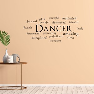May include: A beige wall with a black word cloud that reads "gifted powerful motivated focused graceful dedicated talented flexible DANCER lovely determined persevering amazing disciplined perfectionist strong triumphant". A gold metal table with a round top sits in front of the wall. A vase with a green plant and two decorative objects are on the table.