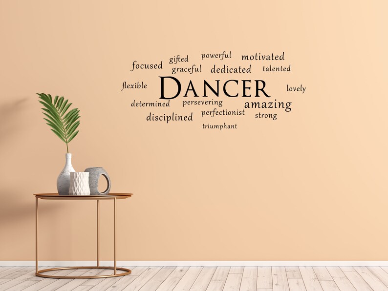 May include: A beige wall with a black word cloud that reads "gifted powerful motivated focused graceful dedicated talented flexible DANCER lovely determined persevering amazing disciplined perfectionist strong triumphant". A gold metal table with a round top sits in front of the wall. A vase with a green plant and two decorative objects are on the table.