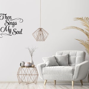 May include: A white living room with a white couch, a gold geometric side table, a dried plant in a vase, and a gold geometric pendant light. The wall has a black vinyl decal with the text "Then Sings My Soul".