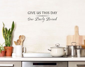 May include: A kitchen scene with a white countertop and a light-colored wall. The image features a potted plant, wooden utensils in a white mug, and stainless steel pots. The text "GIVE US THIS DAY Our Daily Bread" is displayed.
