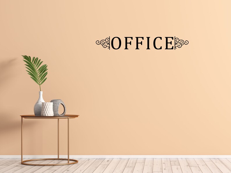 Puede incluir: Una pared de color melocot&oacute;n con la palabra "OFFICE" en letras negras con adornos decorativos a cada lado. Una mesa de metal dorado con tapa redonda se encuentra frente a la pared. Un jarr&oacute;n con una planta verde frondosa y dos piezas de cer&aacute;mica decorativas est&aacute;n sobre la mesa.