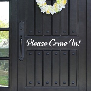 May include: A black front door with a decorative floral wreath and the text "Please Come In!" in white script. The door has vertical panels and decorative hardware.