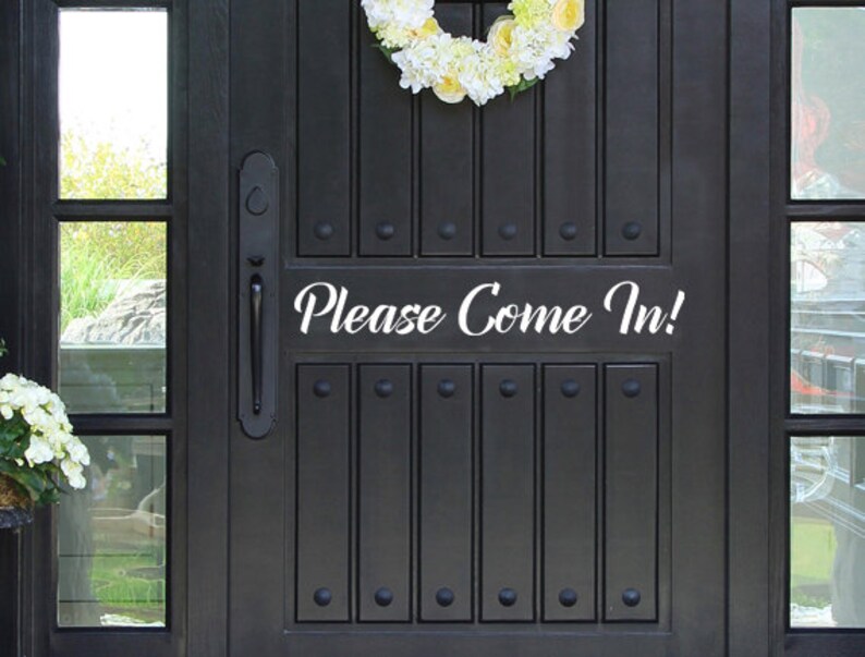 May include: A black front door with a decorative floral wreath and the text "Please Come In!" in white script. The door has vertical panels and decorative hardware.