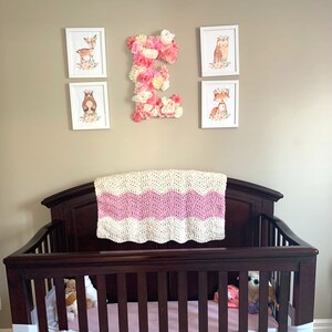 May include: A dark brown wooden cot with a white and pink crocheted blanket on top. The cot has a pink mattress and white safety bumpers. There are stuffed toys inside the cot. The cot is in front of a wall with five framed pictures, including a large floral letter E.