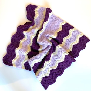 May include: A handmade crochet blanket with a chevron design. The blanket has stripes of deep purple, light purple, and cream. The edges are scalloped, adding a decorative touch to the soft, textured blanket. The blanket is suitable for a bed or sofa.
