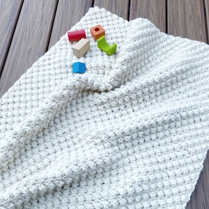 May include: A white crocheted blanket with a textured, bumpy surface.  The blanket is lying on a wooden floor with a few colorful wooden blocks on top.