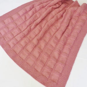May include: A dusty rose-colored knitted blanket with a textured, square pattern. The blanket has a smooth border and is folded, showing the detailed knit design. The soft, warm blanket is suitable for a baby or as a decorative throw.