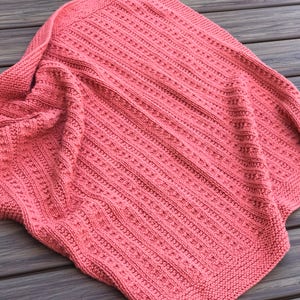 May include: A coral pink knitted blanket with a textured stitch pattern. The blanket is folded and draped over a wooden surface.