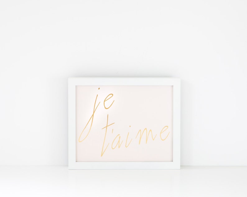 Puede incluir: Marco de imagen blanco con fondo rosa claro y letras doradas que dicen "je t'aime".