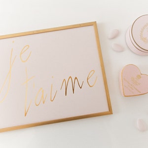 Puede incluir: Un cuadro con marco dorado con fondo rosa y las palabras "je t'aime" escritas en letras doradas. Hay una caja rosa en forma de coraz&oacute;n y una caja redonda rosa con ribetes dorados y un dise&ntilde;o floral sobre una superficie blanca.