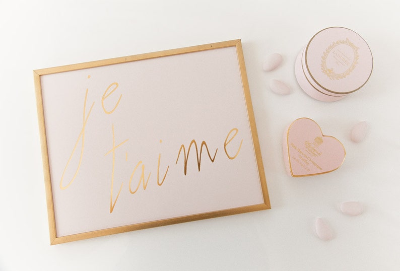 Puede incluir: Un cuadro con marco dorado con fondo rosa y las palabras "je t'aime" escritas en letras doradas. Hay una caja rosa en forma de coraz&oacute;n y una caja redonda rosa con ribetes dorados y un dise&ntilde;o floral sobre una superficie blanca.