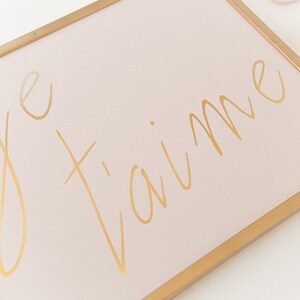 Puede incluir: Un lienzo rosa enmarcado con letras doradas que dicen "je t'aime".