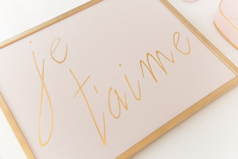 Puede incluir: Un lienzo rosa enmarcado con letras doradas que dicen "je t'aime".