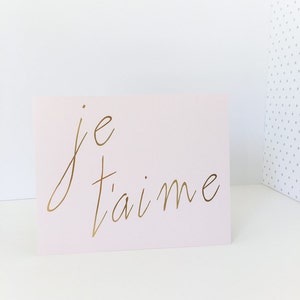 Puede incluir: Una tarjeta rosa con las palabras "je t'aime" escritas en letras doradas.