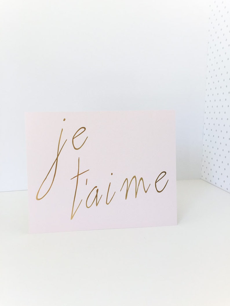 Puede incluir: Una tarjeta rosa con las palabras "je t'aime" escritas en letras doradas.