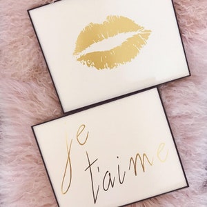Puede incluir: Dos impresiones enmarcadas con letras doradas. La impresi&oacute;n superior presenta una impresi&oacute;n de labios de brillo dorado. La impresi&oacute;n inferior dice "Je t'aime" en letras de brillo dorado.