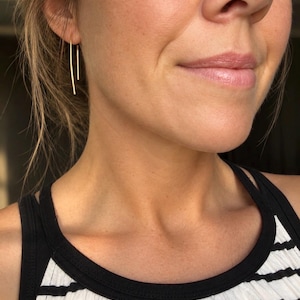 May include: A pair of gold-toned threader earrings featuring a thin, curved wire design that hangs vertically from the earlobe. The earrings consist of a single continuous piece of metal that loops through the piercing, creating two parallel lines.