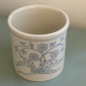 May include: A cylindrical, off-white ceramic crock with a blue floral design. The design features stylized flowers and leaves. The crock has a rounded rim and a smooth, unglazed surface. It is a vintage-style kitchen or home decor item.