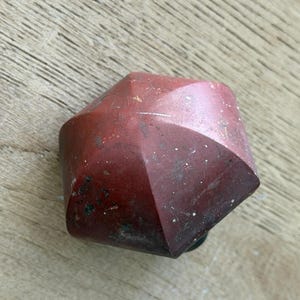 May include: A faceted, geometric object with a reddish-brown color and a slightly weathered appearance. The object has a dark base and appears to be made of a solid material. The surface shows subtle variations in color and texture.