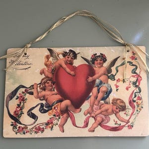 May include: Vintage Valentine's Day sign featuring cherubs, a large red heart, and floral ribbons. The sign has a cream-colored background with the text "Be my Valentine." A gold ribbon is attached for hanging.