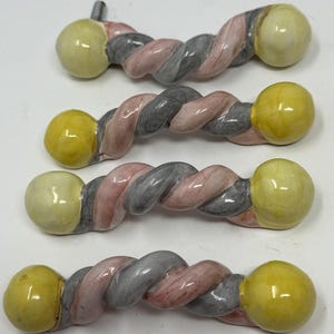 May include: Four ceramic cabinet pulls. Each pull features a twisted design in pink, gray, and white, with a yellow sphere at each end. One pull has a metal screw attached. These decorative pulls are for furniture.