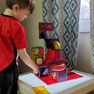May include: A child plays with a colorful magnetic tile construction set. The set includes clear, translucent tiles in various shapes and sizes, as well as tubes and connectors. The tiles are arranged to create a structure with a ramp and a ball run.