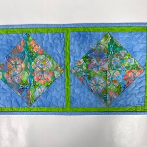 May include: A quilted textile with a light blue background and two large diamond shapes. The diamonds feature a floral pattern in shades of green, orange, and purple. A green border frames the diamonds and the edges of the textile.