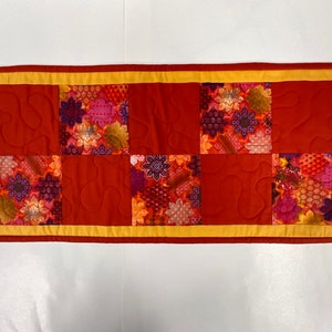 May include: A red quilted table runner with a yellow border. The runner is made of fabric with a floral pattern in shades of red, orange, and purple.