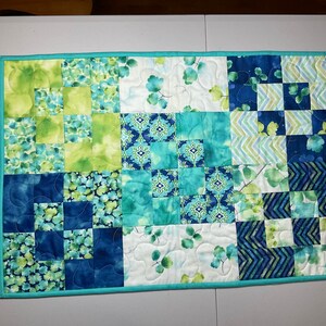 May include: A blue and green patchwork quilt with a teal border. The quilt is made of various fabrics with different patterns, including leaves, flowers, and geometric designs.