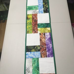 May include: A green and white table runner with a geometric pattern of colorful fabric squares. The runner is made of patchwork fabric with floral and abstract designs.