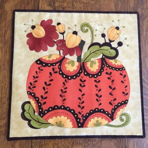 May include: A quilted fabric wall hanging featuring a red and orange pumpkin with black and white accents. The pumpkin is surrounded by yellow and red flowers with green leaves.