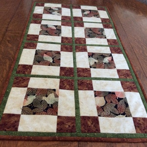 May include: A brown and green checkered table runner with a leaf pattern. The runner is made of fabric and has a quilted texture.