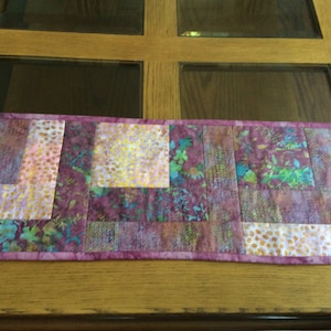 May include: A purple, pink, and gold patchwork table runner with a floral pattern. The runner is made of fabric squares sewn together.