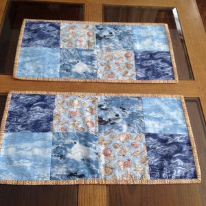 May include: Two blue and white quilted table runners with a seashell pattern. The runners have a tan fringe border.