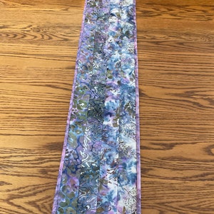 May include: A purple and blue table runner with a floral pattern. The runner is made of fabric and has a quilted texture.