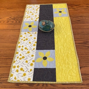 May include: A yellow, gray, and brown quilted table runner with a floral pattern. The runner has a yellow border and is decorated with yellow flowers. A small green bowl is placed on the runner.