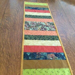 May include: A multi-colored table runner with a patchwork design. The runner features stripes of green, brown, orange, yellow, and blue fabric. The edges are trimmed with a gold fabric.